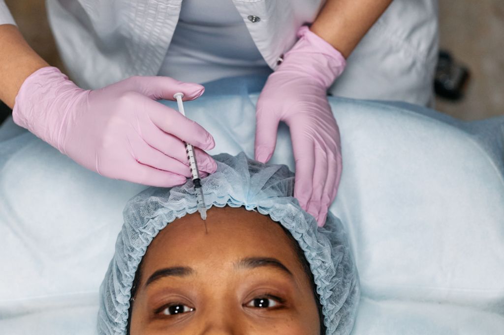 Close-up photo of a Black woman laying on her back, receiving Botox injections in her forehead from a medical professional.